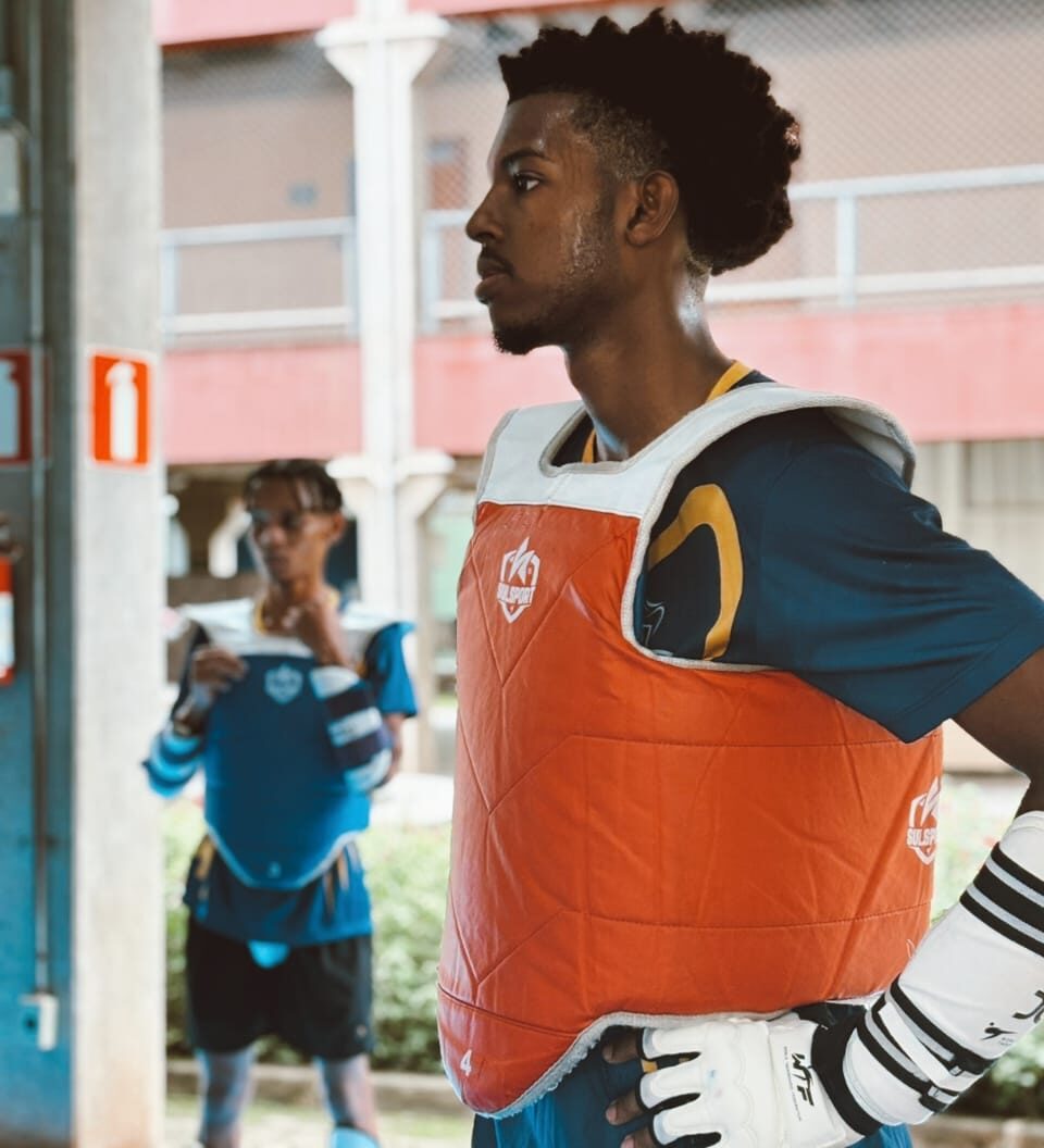 Em maio, o jovem Humberto Vinicius, de 19 anos, deixou o bairro Fênix, em Itabira, rumo ao Rio de Janeiro para disputar o Pan-Americano de Taekwondo. E não foi em vão. Na disputa ocorrida entre 6 e 10 de maio, o atleta conquistou o ouro. Mas ele quer mais. Em entrevista à DeFato, Humberto contou a sua história e dividiu seu grande sonho: chegar à elite mundial do esporte que transformou inquietude em competição. “Conheci o taekwondo pelo projeto social Crianças do Amanhã, antes localizado no bairro João XXIII e hoje no Bálsamos. Desde pequeno, sempre gostei muito de ação, assistia filmes de luta com meu pai. Gostava muito do incrível Hulk, da agressividade dele, queria ser ele”, lembra. Atleta desde os 15 anos, Humberto conheceu o taekwondo após se aventurar em outras áreas. Não deu certo. A verdadeira paixão seria descoberta aos 8 anos, com uma inspiração caseira. “Meu irmão mais velho sempre foi um exemplo pra mim. Ele praticava muitos esportes por lazer, mas, ainda assim, quase chegou à faixa preta, mas teve que parar pra trabalhar. Ele me mostrava as faixas, medalhas, fotos e eu achava incrível. Meu pai primeiro me colocou no karatê, não gostei. Depois fiz uma aula de capoeira, também não gostei. Mas com o taekwondo foi amor à primeira vista”. A partir dali, Humberto aprendeu não só a dominar o perfil agitado, como também a se descobrir como pessoa. “O esporte, para mim, significa um estilo de vida. O taekwondo, desde a infância, me ensinou coisas como disciplina, integridade, autocontrole, espírito indomável. E também foi essencial para meu crescimento, minha formação como pessoa, caráter e princípios. Sem contar a saúde física, fiquei muito alto pela prática de esportes. Já cheguei a fazer, ao mesmo tempo, taekwondo, futsal e futebol de campo. Então sempre gostei muito de esporte e para mim é um estilo de vida que agrega muito física e mentalmente”, avalia. Presente e futuro Sobre a recente vitória, o taekwondista confessa ainda estar a assimilando. Ele também relembra outra participação na mesma competição há três anos. “Ainda estou processando a conquista. Foi minha segunda disputa de Pan-Americano, o primeiro foi na República Dominicana em 2023, estava com a seleção brasileira. E como eu tinha participado daquela vez, eu já tinha um pouco de noção do nível dos atletas. O pan do Rio era aberto, não seriam obrigatoriamente todos os países da América. Mas havia atletas de ponta, apenas cabeças de chave”. Aos poucos, a ansiedade foi dando lugar à confiança na busca por uma medalha que, para Humberto, era uma meta inegociável. “Fui confiante, mas com certo receio, porque eu tinha me arriscado a participar deste campeonato mesmo sem ter condições financeiras. Pedi ajuda a amigos e familiares e consegui ir, mas pensando ‘preciso sair daqui com uma medalha’. Estudei as lutas e os adversários e à medida que as lutas foram passando e eu fui percebendo que estava mais perto do ouro, a realidade meio que se distorceu”, descreve. “Sair do bairro Fênix, em Itabira, para ir ao Rio de Janeiro na Arena Carioca - onde aconteceram as Olimpíadas de 2016 -, pisar no tatame e levar a medalha de ouro para mim é coisa de filme”. Embora gigante, o feito não deixa Humberto em uma zona de conforto. O jovem itabirano agora deseja ser reconhecido fora do país e marcar seu nome na história do esporte. “Meu maior sonho é chegar o mais longe possível. Quero chegar aos campeonatos internacionais e ter meu merecido reconhecimento. Já fui atleta da seleção duas vezes, ganhei campeonatos nacionais e até então não fui reconhecido como merecia. Não quero desistir desse sonho por questões financeiras. Desejo chegar nas Olimpíadas e não apenas estar lá, mas ser o melhor. Quero que as pessoas, mesmo depois que eu me aposente, lembrem de mim e pensem ‘não vai haver um atleta como o Humberto foi’”, finaliza.